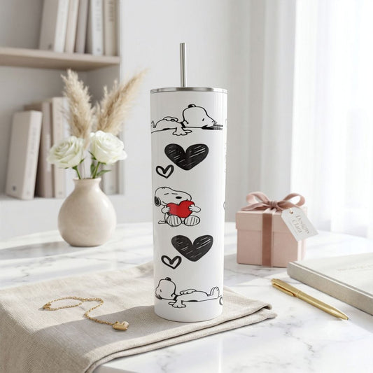 A tall white insulated tumbler with a stainless steel straw, printed with black heart shapes and Snoopy illustrations, standing on a beige cloth on a marble table in a bright room.